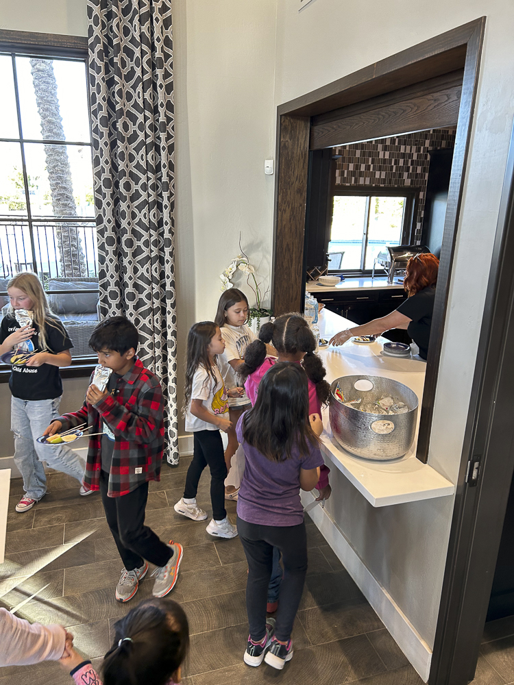 Children gather at a serving counter, eagerly receiving food from a server. A large silver bowl is filled with snacks. The room has patterned curtains and wooden flooring. Large windows provide natural light.