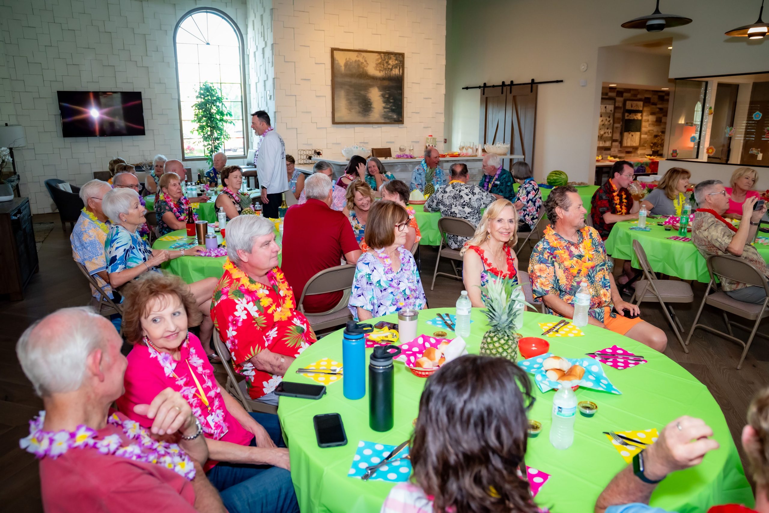 A group of people in colorful Hawaiian shirts and leis sit around tables with green tablecloths and tropical decorations, attending a lively indoor gathering in a bright, spacious room.