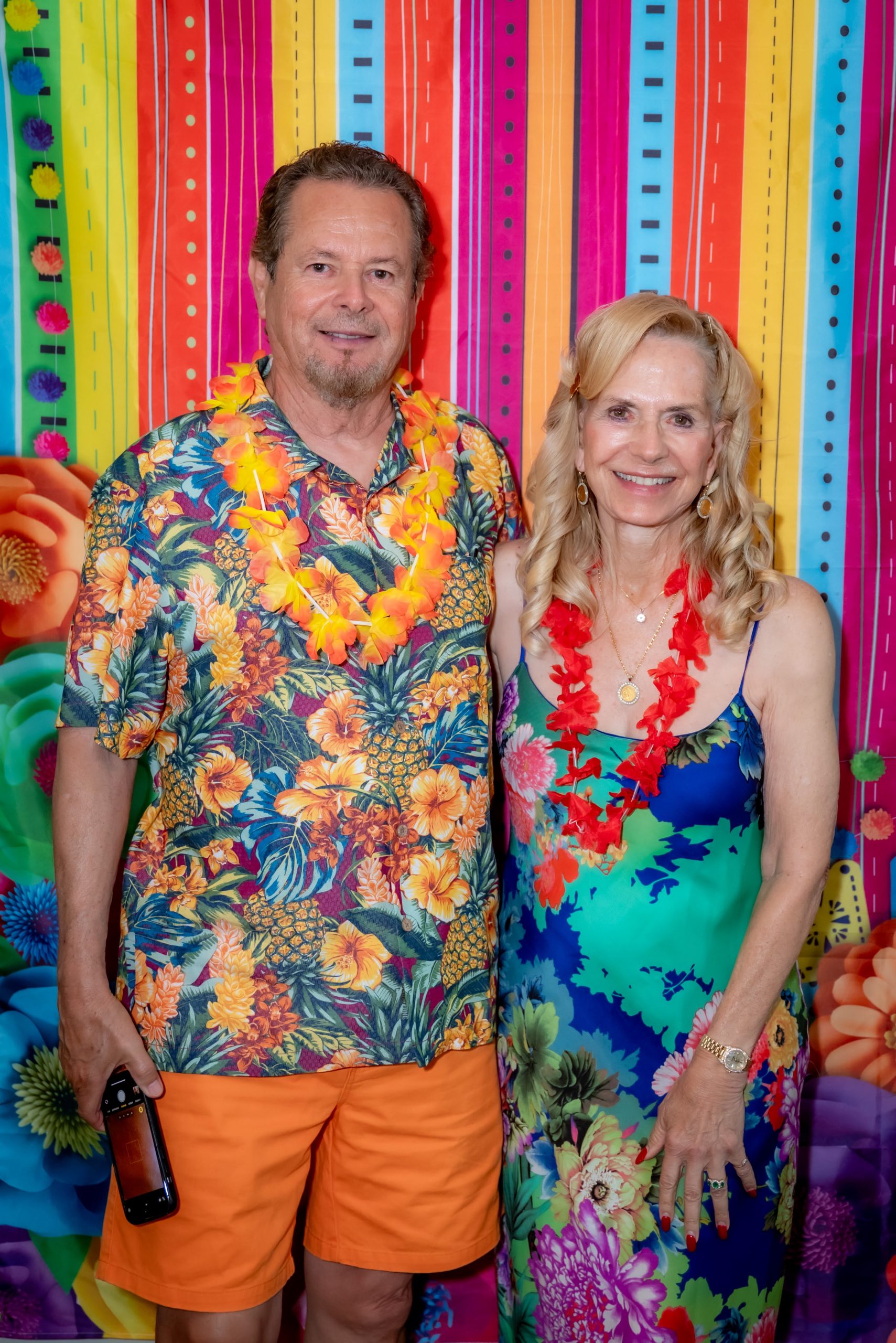 A man and woman in colorful, tropical-themed outfits and leis stand smiling in front of a bright, multicolored floral and striped backdrop. The man holds sunglasses and wears orange shorts; the woman wears a floral dress.