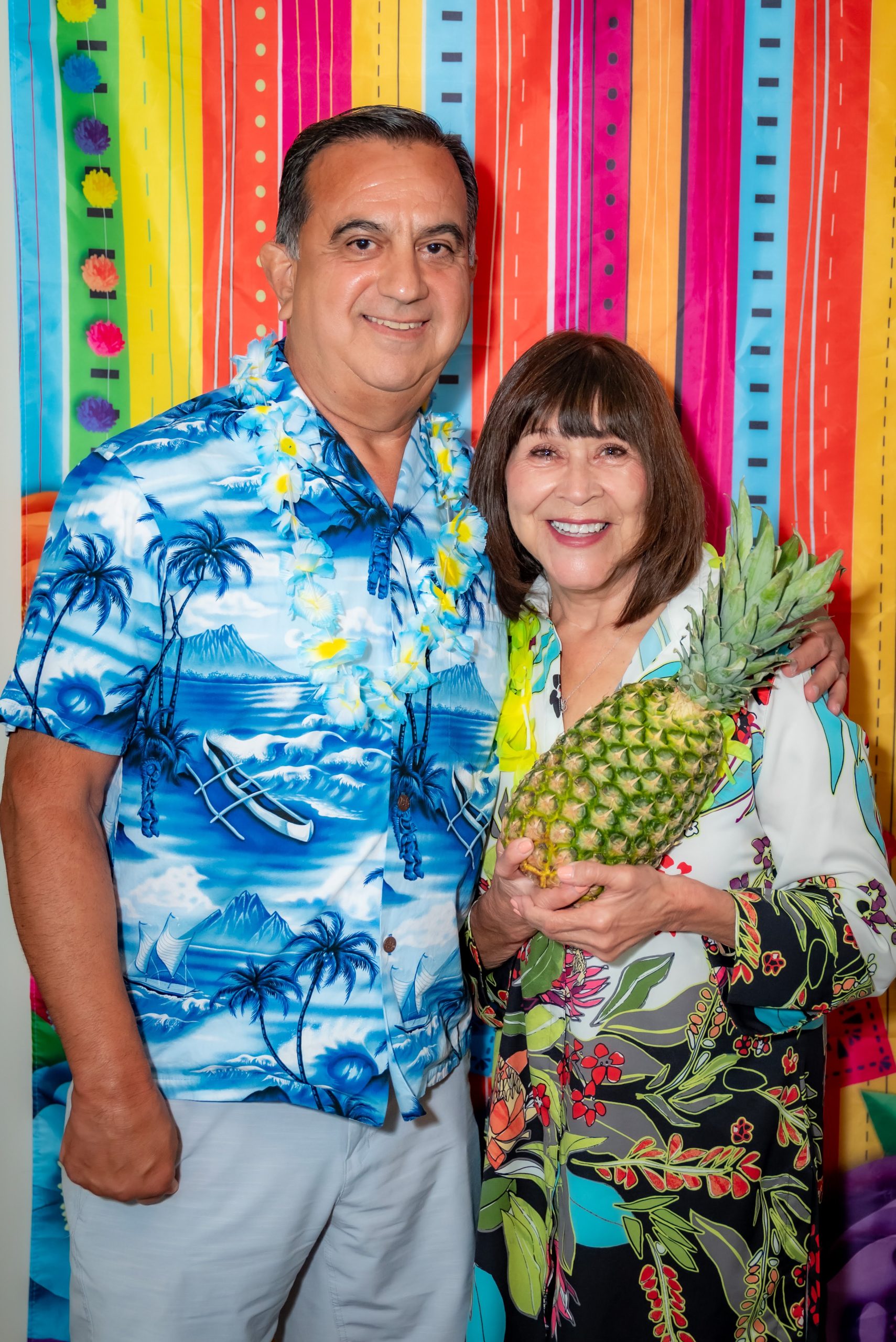 A smiling man in a blue Hawaiian shirt and a lei stands next to a woman in a floral dress holding a pineapple, with a colorful striped backdrop behind them.