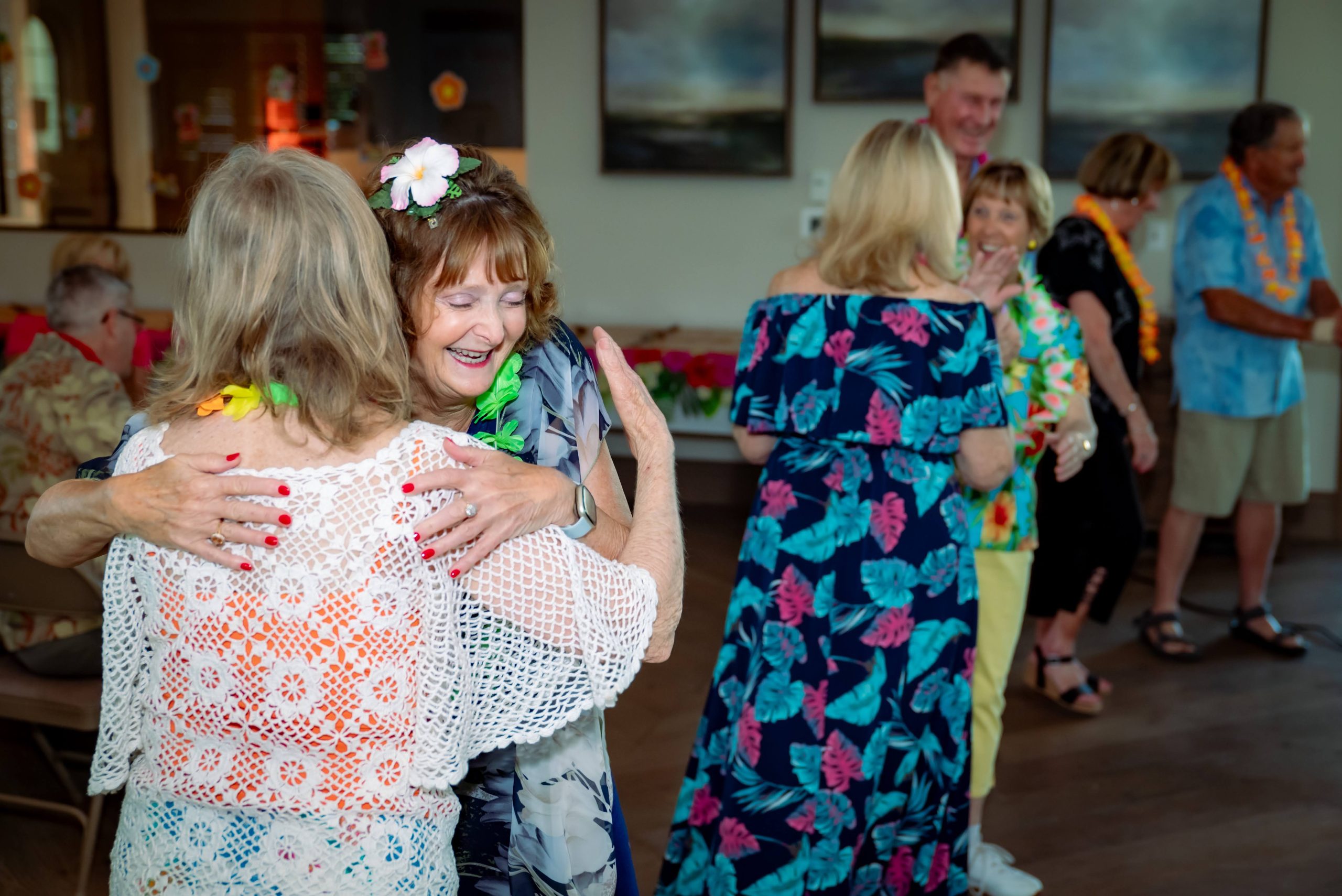 Two women wearing floral leis and summer clothes hug and smile at a lively indoor gathering, while other people in colorful outfits socialize in the background.