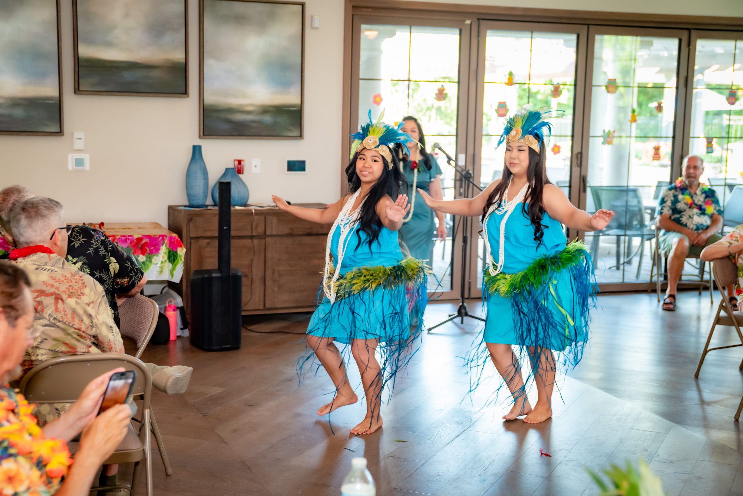 Two women in blue dresses with grass skirts and flower headpieces perform a traditional Hawaiian dance in a bright room, while an audience watches and takes photos.