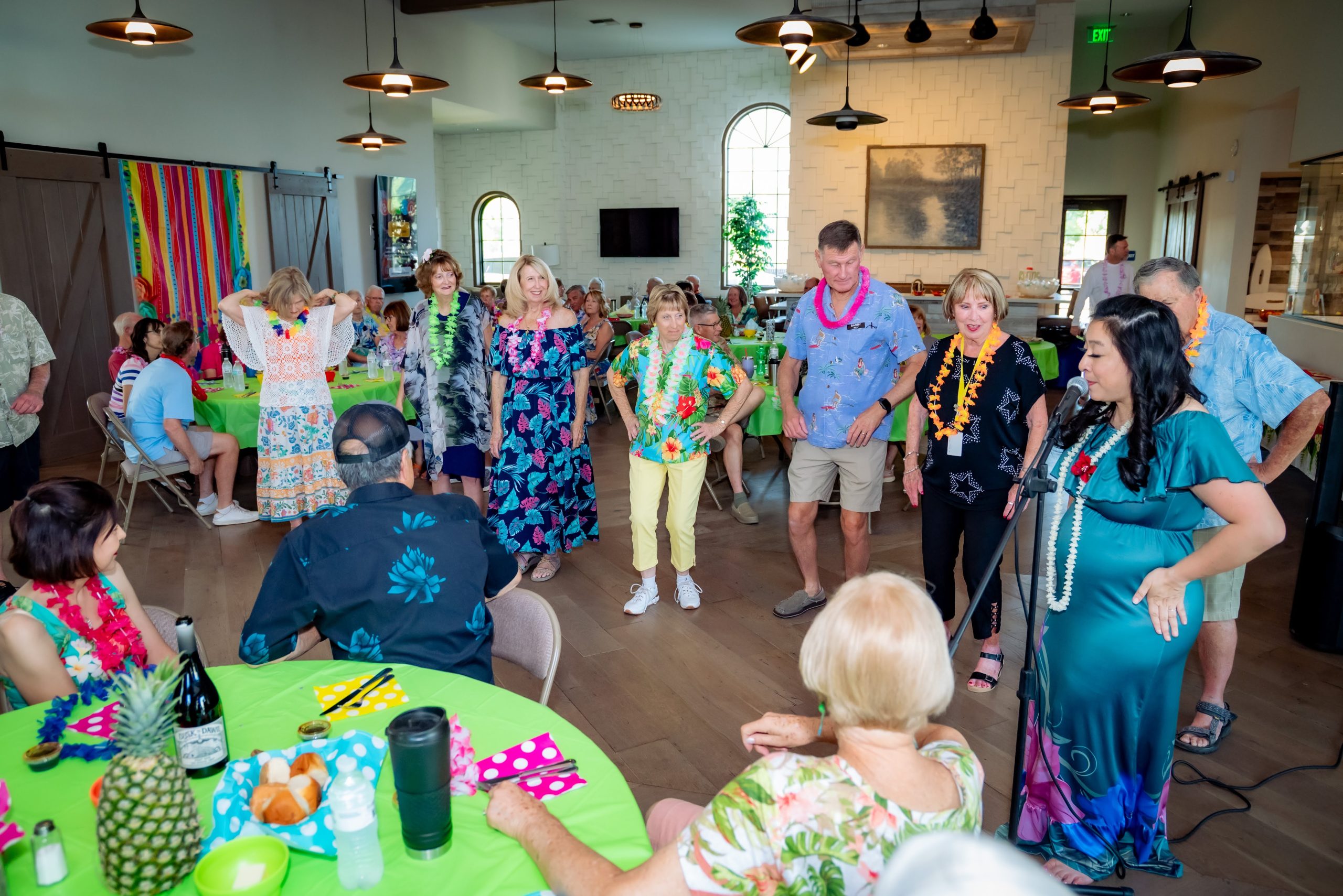 A group of people in colorful, tropical-themed clothes and leis gather in a decorated room with round tables, chatting and standing near the center, while others sit and eat. Bright decorations and fruit centerpieces set a festive mood.