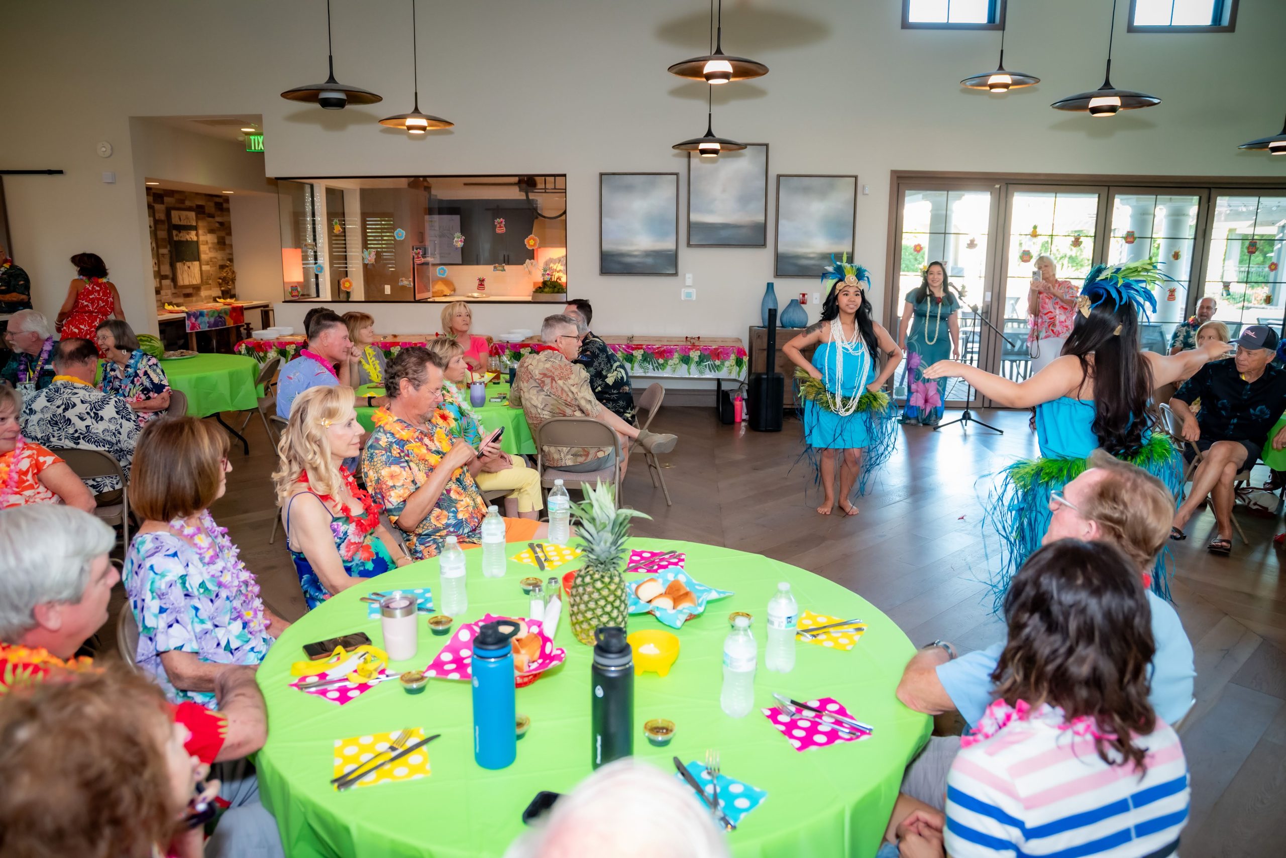 A group of people in colorful Hawaiian shirts watch dancers in grass skirts and headdresses perform at a luau-themed indoor party with tropical decorations and vibrant table settings.