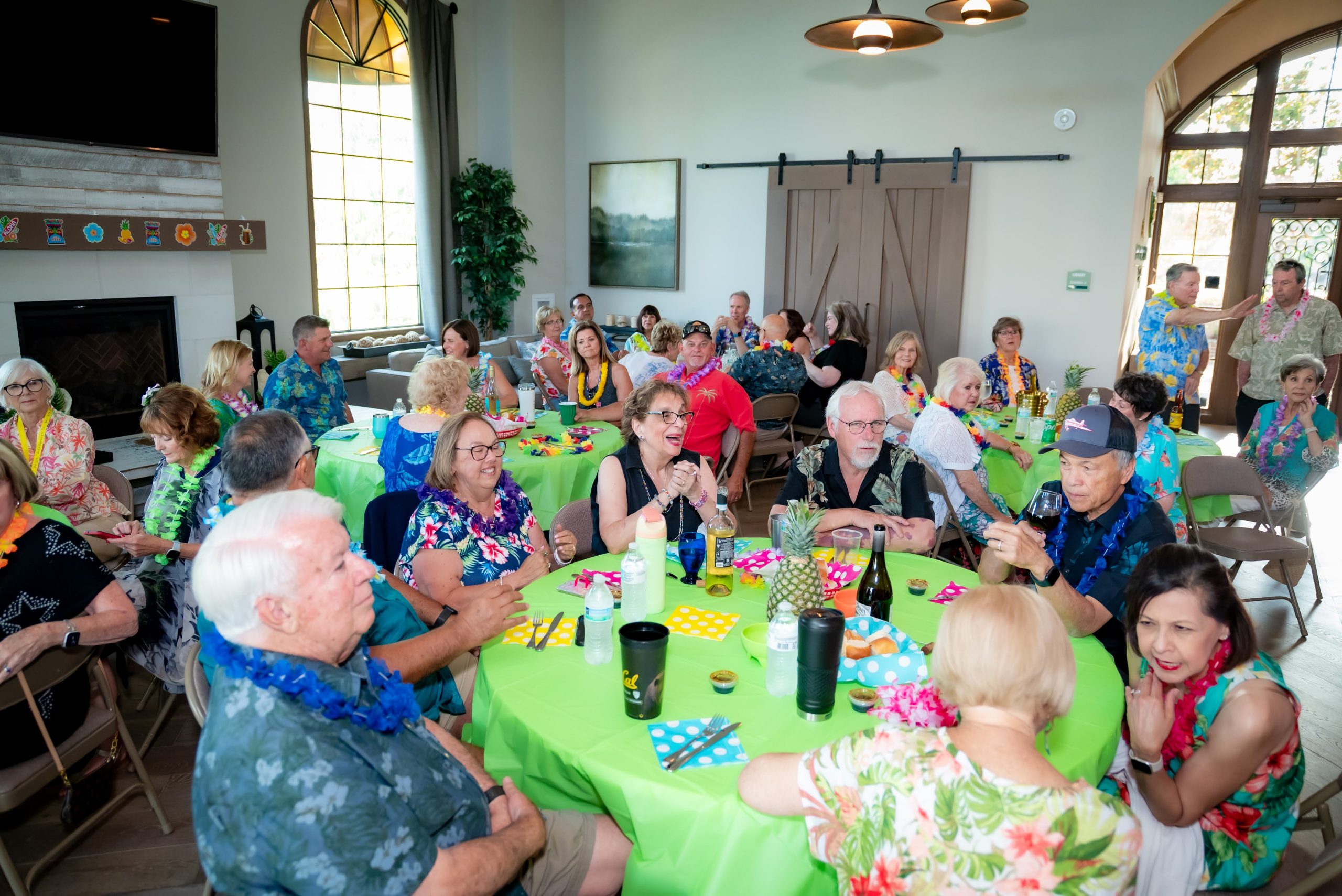A group of older adults, many wearing Hawaiian shirts and leis, sit around decorated green tables at a lively indoor party. Some are laughing, talking, or holding drinks in a bright, spacious room with large windows.