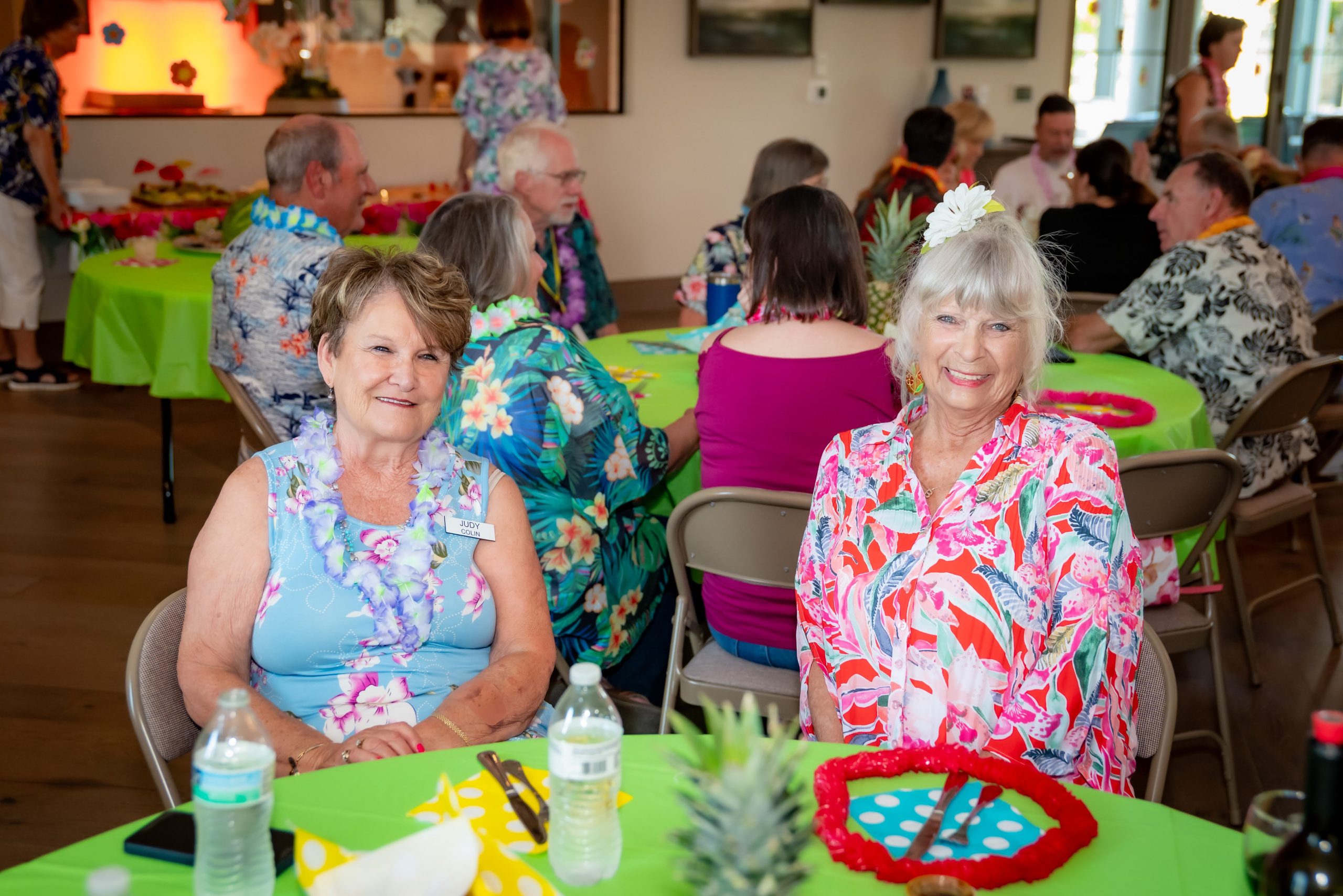 Two older women in colorful Hawaiian shirts smile at a table decorated with green cloth and tropical items at a lively indoor party with other people in similar attire in the background.