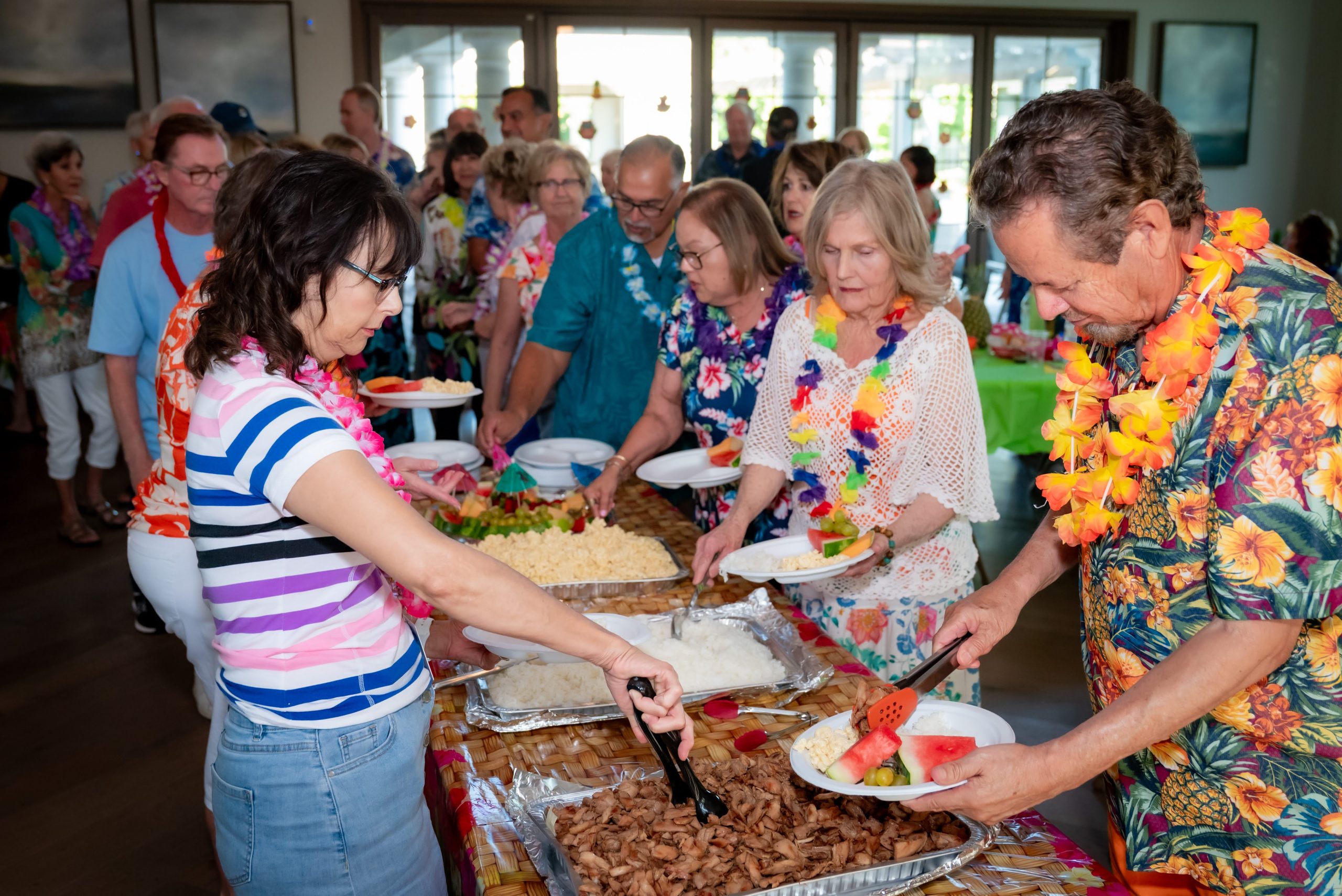 A group of people wearing colorful leis and Hawaiian shirts serve themselves food from a buffet table at a lively indoor gathering. Plates are being filled with fruit, salad, and cooked dishes.