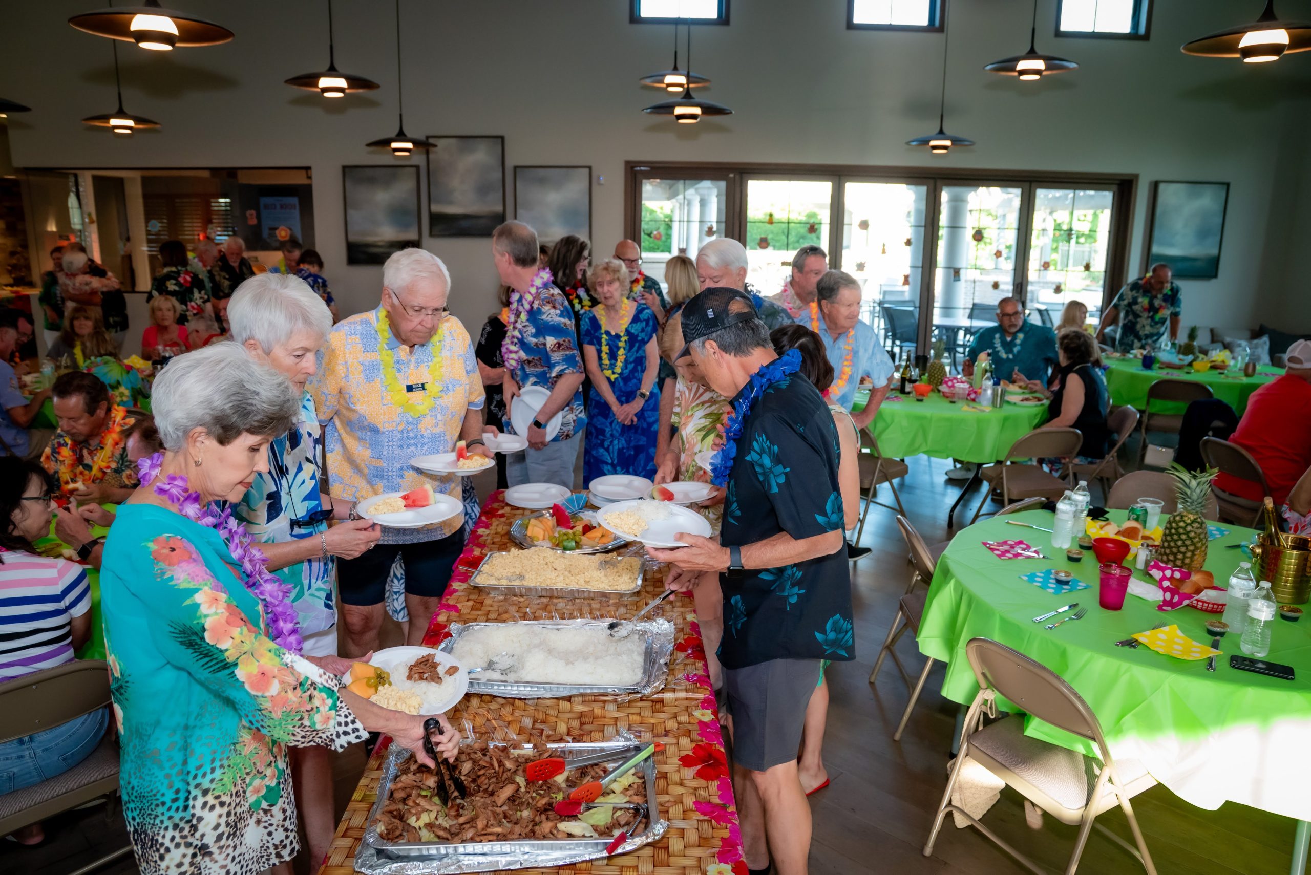 A group of people in colorful Hawaiian shirts serve themselves food from a buffet table at a festive indoor gathering with round tables, decorations, and natural light.