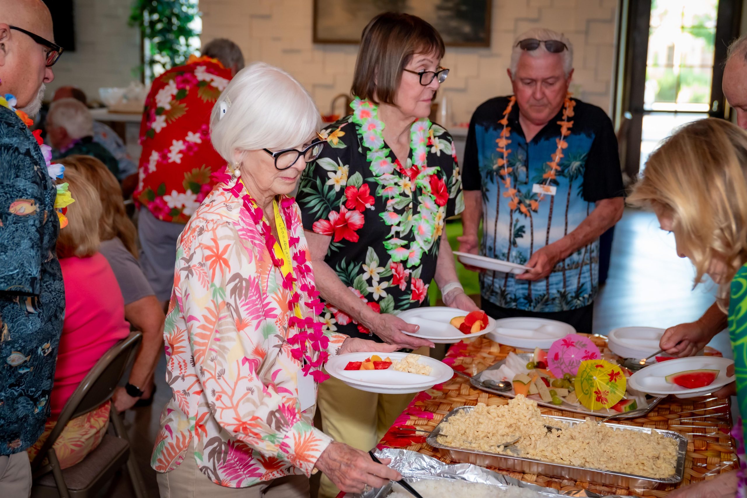 A group of older adults in colorful Hawaiian shirts and leis serve themselves food from a buffet table, with plates of rice and fruit, at a lively indoor gathering.
