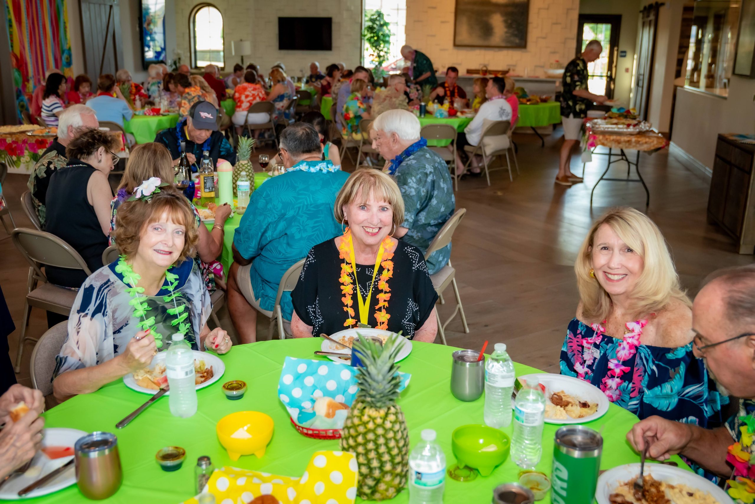A group of people enjoy a festive meal at a tropical-themed party, wearing colorful leis and floral outfits, seated around decorated tables with food, drinks, and pineapples. The room is lively and filled with conversation.