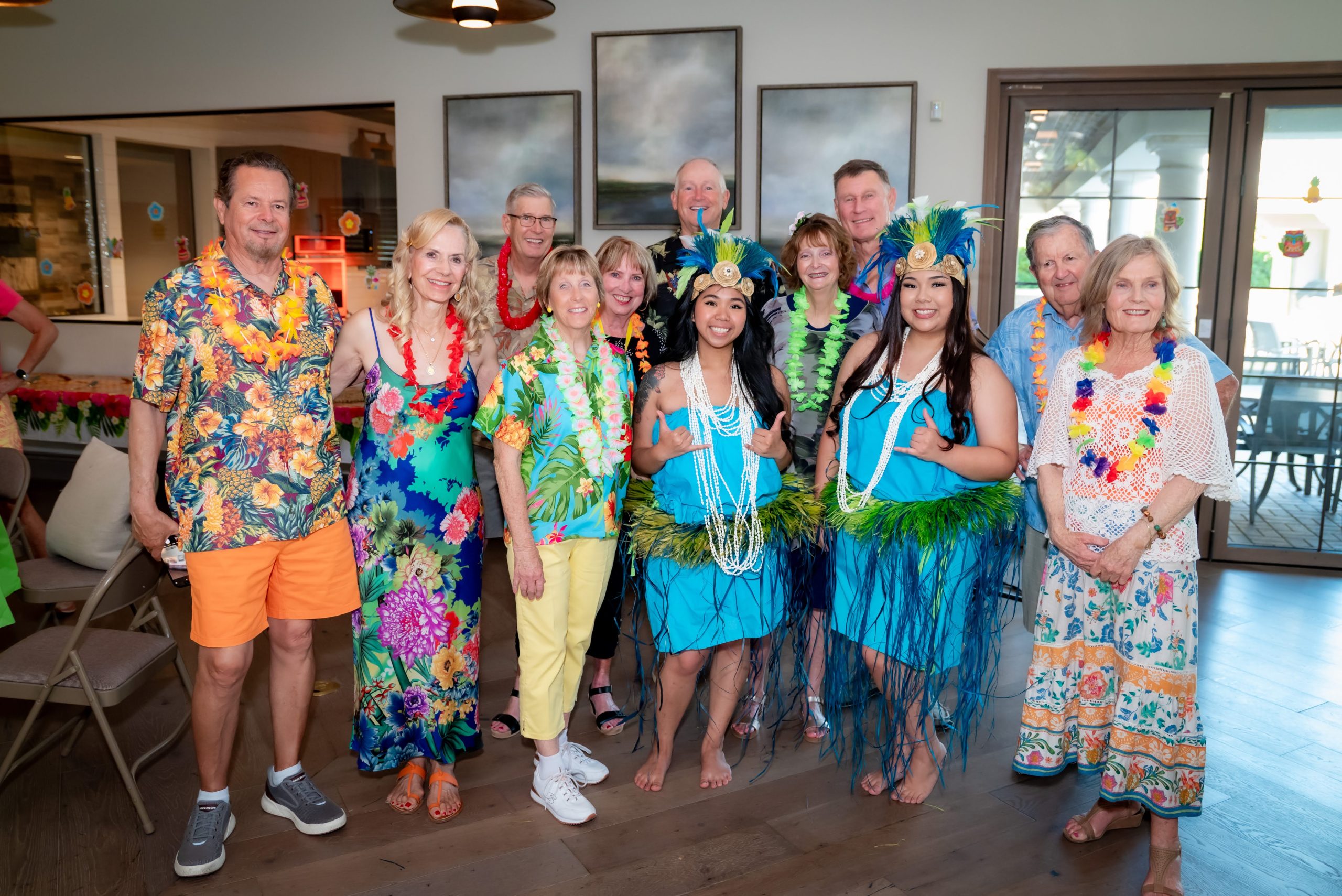 A group of people pose indoors wearing colorful Hawaiian-themed outfits and leis. Two women in blue hula costumes with grass skirts and flower crowns stand in front, smiling with others in festive, tropical attire behind them.