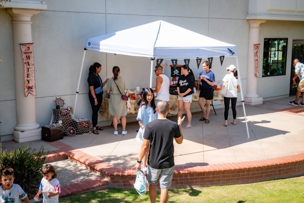 A small group of people gather under a white canopy tent decorated with pirate flags outside a building. Some are serving snacks on tables while others chat nearby; a few children and adults walk on the grass in front.