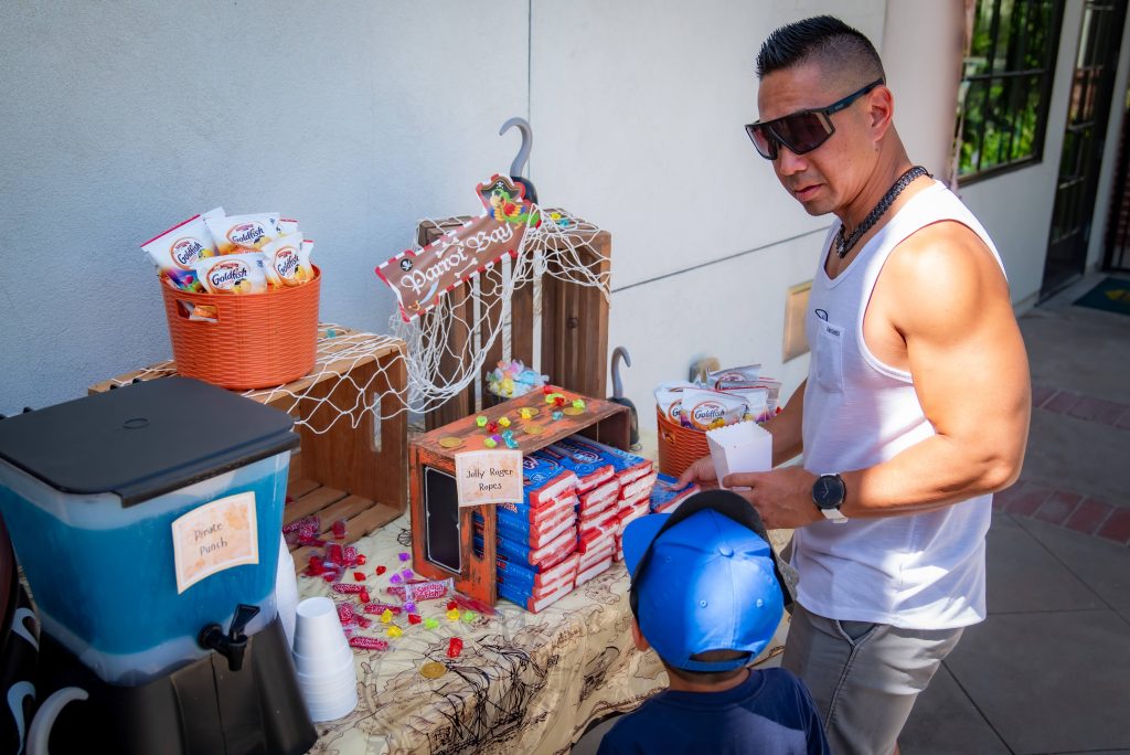 A man in sunglasses and a white tank top stands with a child at a decorated table with drinks, snacks, and candy displayed in baskets and boxes outdoors.