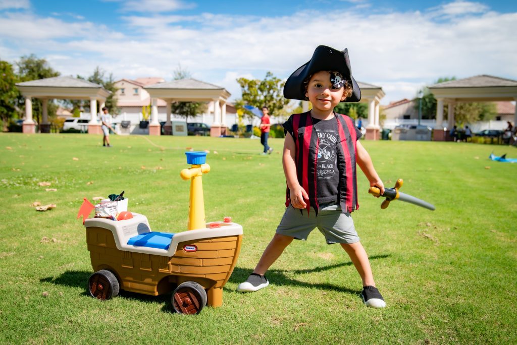 A young child dressed as a pirate, wearing a hat and an eye patch, smiles while holding a toy sword beside a toy pirate ship on a grassy field in a park, with gazebos and houses in the background.
