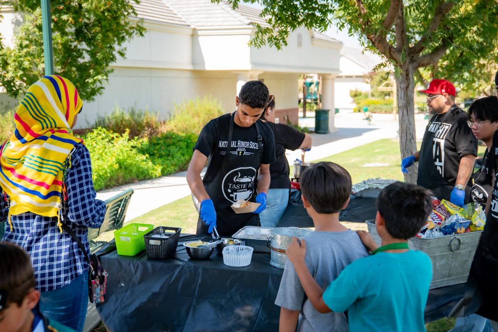 A man serves food to children and adults from trays on a table covered with a black cloth at an outdoor event, with trees and a building in the background.