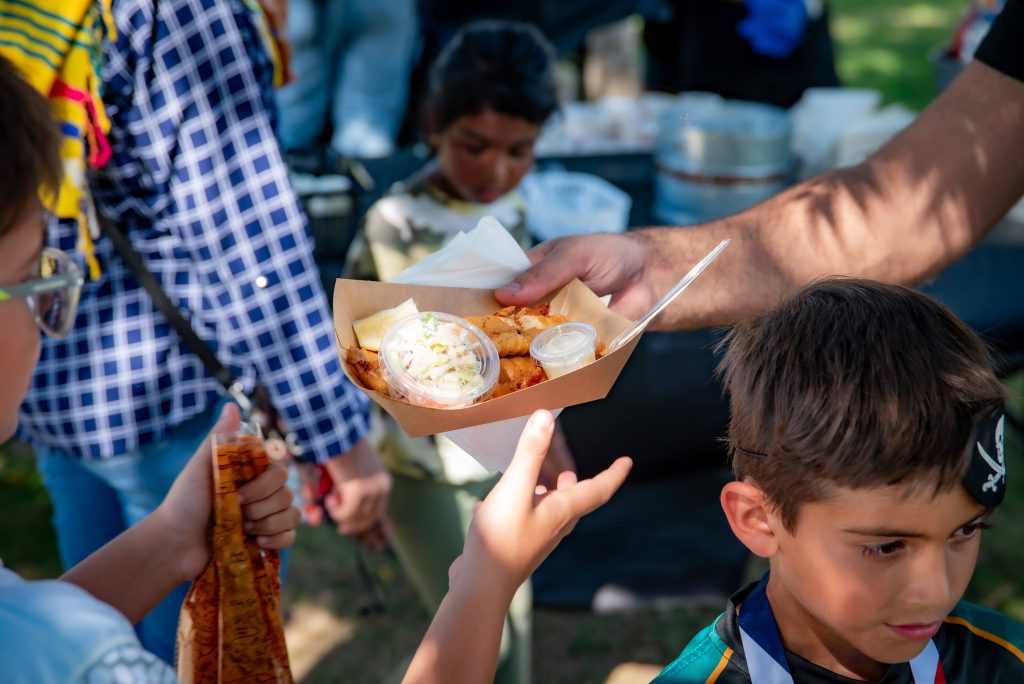 A person hands a tray of food, including fries and coleslaw, to a child at an outdoor event. Other children and adults are nearby, with blurred backgrounds suggesting a busy, festive atmosphere.