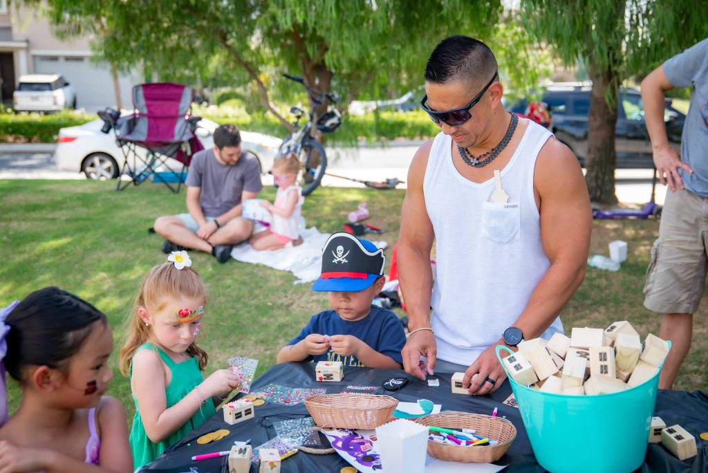 A man helps three children with arts and crafts at an outdoor table. The kids wear face paint and pirate hats. Other people relax on the grass in the background on a sunny day.