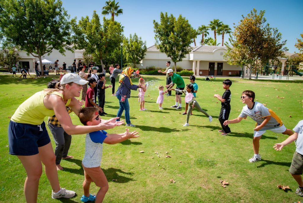 Children and adults play an outdoor game on a sunny day in a grassy park, forming lines and tossing objects to each other, surrounded by trees and buildings in the background.