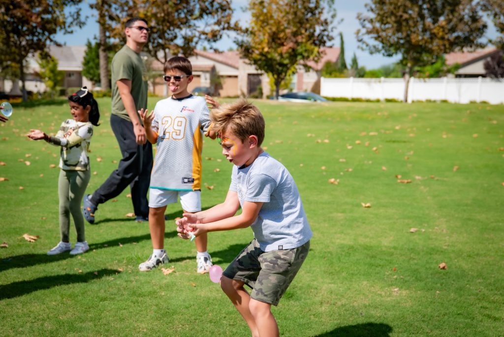 Children play a game outside on a grassy field, concentrating as they handle water balloons. Trees and houses are visible in the background on a sunny day.