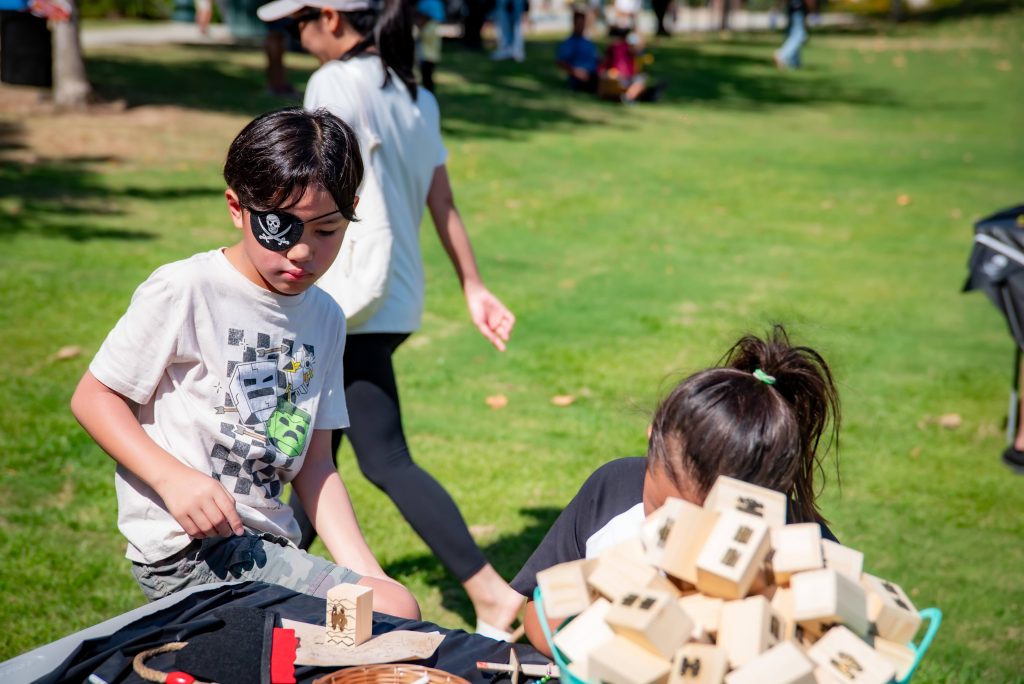 A boy wearing a pirate eye patch plays with wooden blocks at a table outdoors, while a girl sits nearby with more blocks. Other people walk on the grass in the background on a sunny day.