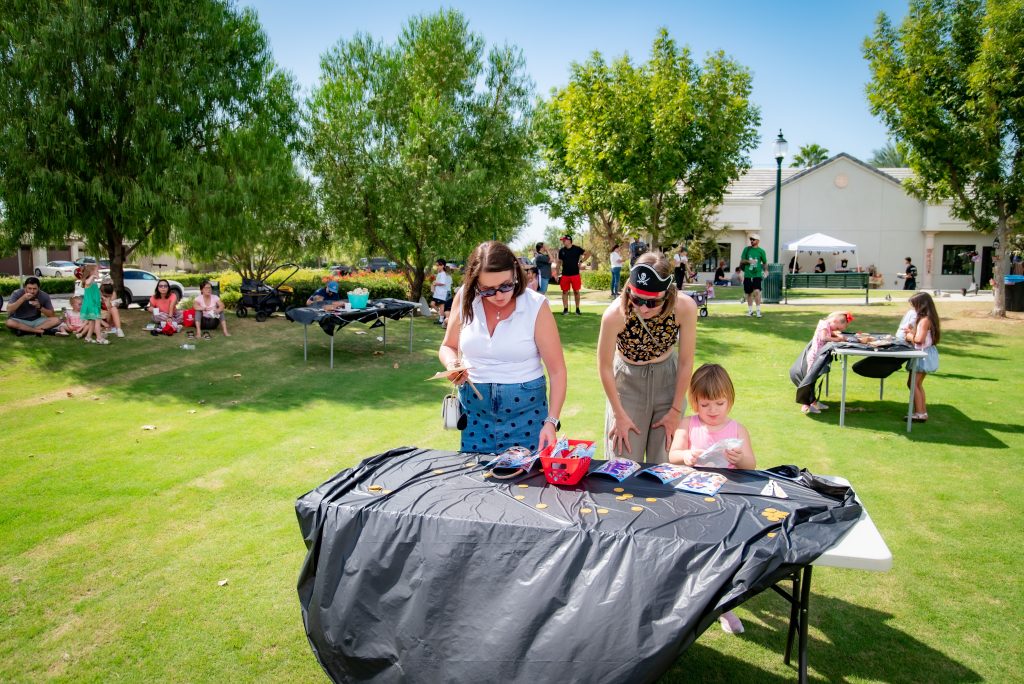 Two women and a young girl work on crafts at a table covered with a black cloth in a sunny park, with other people and groups sitting and standing on the grass in the background.