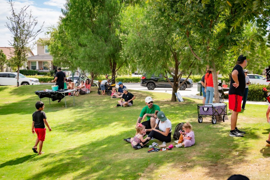 Families and children are relaxing on a grassy area in a park, sitting in small groups under trees. Kids play with toys and adults chat. Cars are parked nearby and houses are visible in the background.