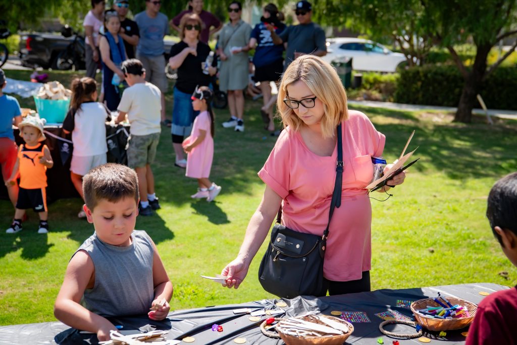 A woman and a young boy work on crafts at an outdoor table during a community event, with other people and children gathered in the background on a sunny day.