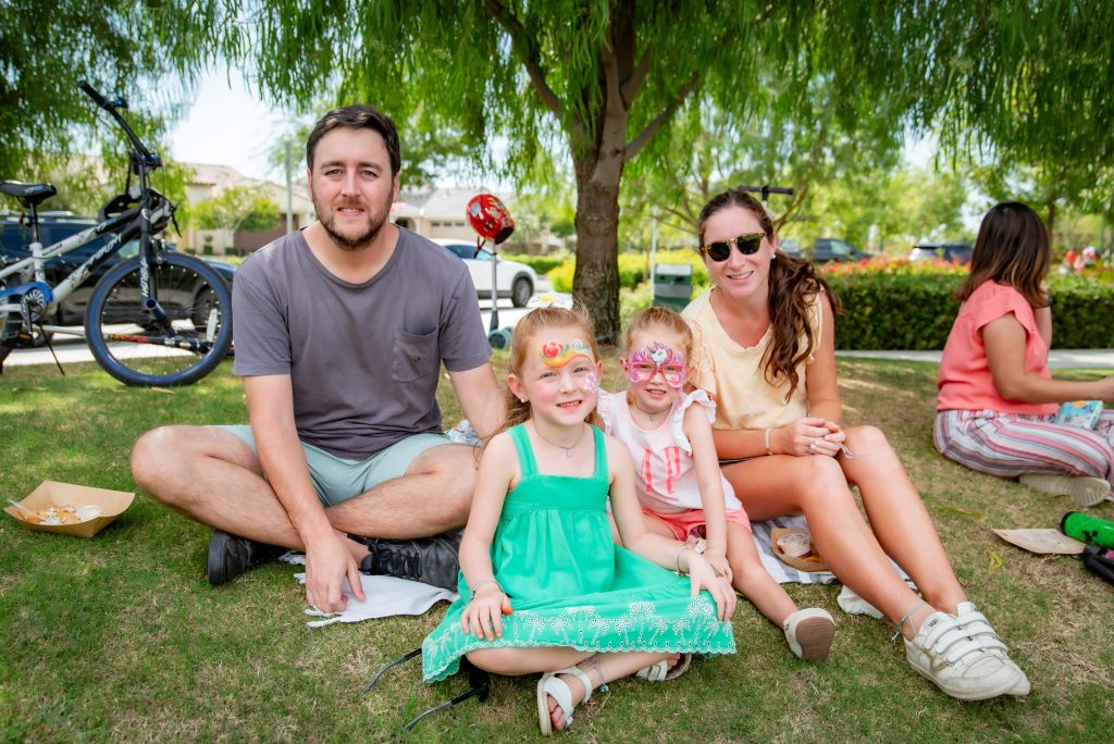 A family of four sits on the grass under a tree at a park. The parents smile at the camera while two young girls with face paint sit between them. A bicycle and other people are in the background.