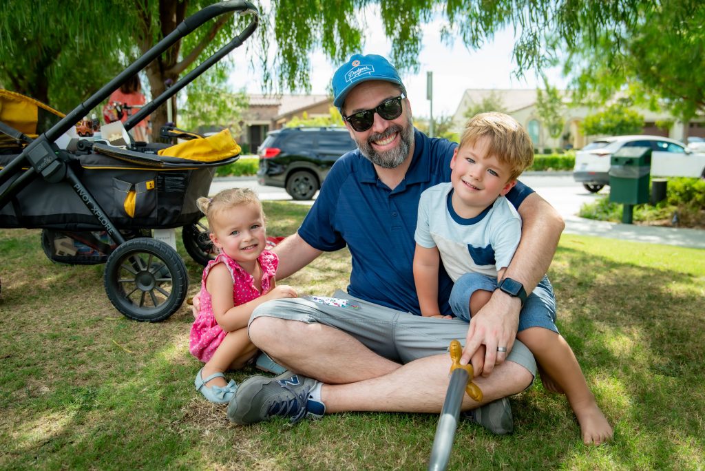 A smiling man in sunglasses and a blue cap sits on the grass with two young children, a girl in a pink dress and a boy in a blue shirt, beside a stroller in a sunny park.