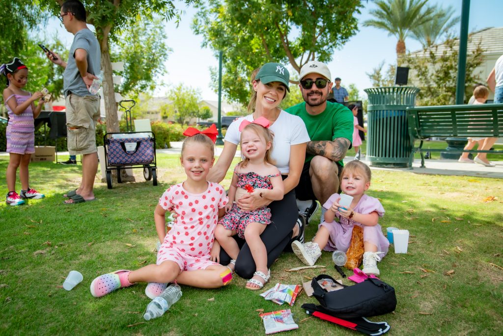 A smiling family with three young children sits on grass at a park, surrounded by snacks and drinks, on a sunny day. Adults and children are seen in the background enjoying the outdoor setting.
