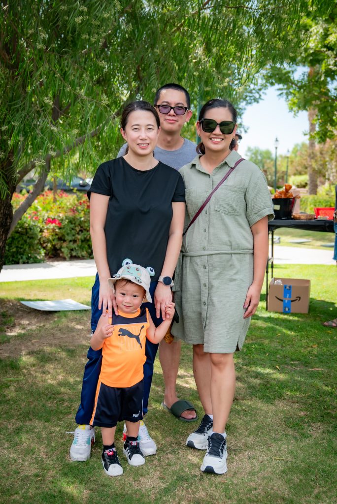 Four adults and a young child stand together outdoors on grass, smiling at the camera. The child wears an orange outfit and hat. Trees and park scenery are in the background on a sunny day.