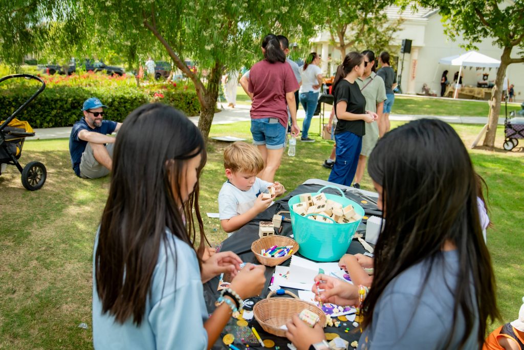 Children are gathered around a table outdoors, making crafts with wooden blocks and art supplies. Adults and other children are nearby, some standing and some sitting on the grass under trees on a sunny day.