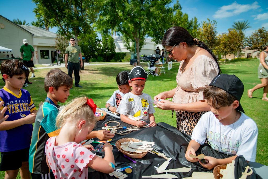 A group of children, some wearing pirate hats, gather around a table outdoors doing arts and crafts with a woman assisting them. The scene is set on a grassy field with trees and buildings in the background.