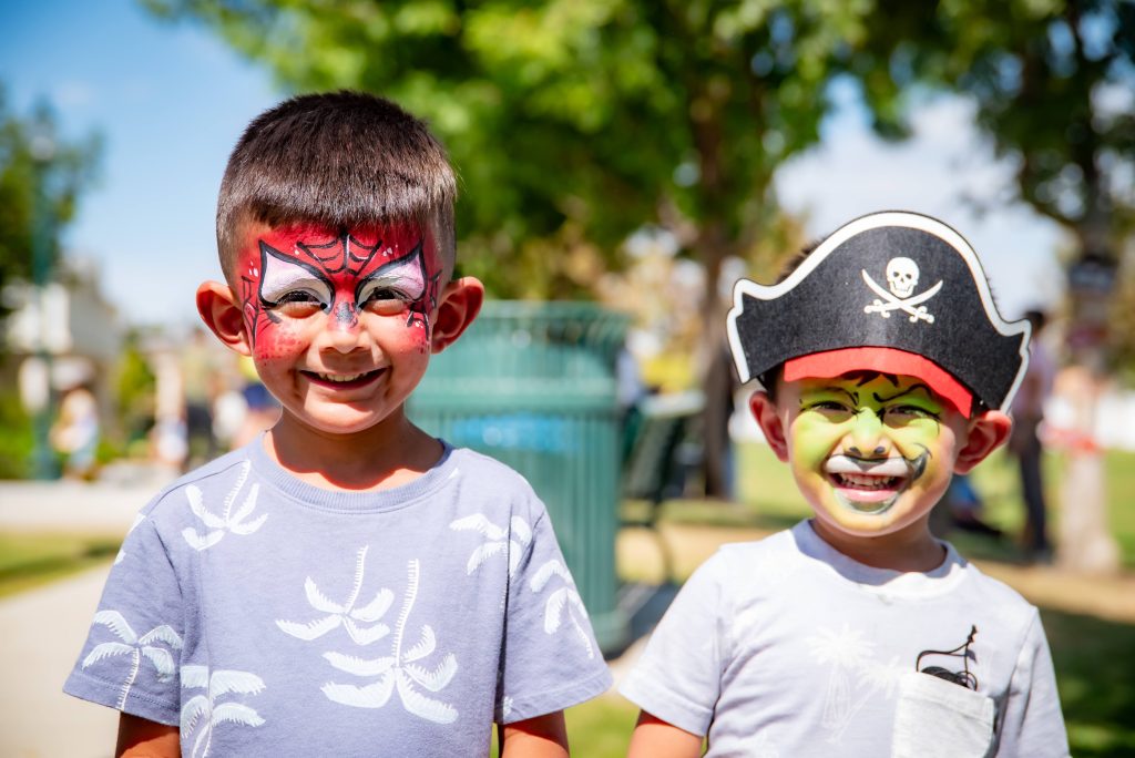 Two young boys smile outdoors. One has a Spiderman face paint, the other has a pirate hat and green face paint. Both wear light blue shirts with white patterns, and the background is a sunny park with trees.