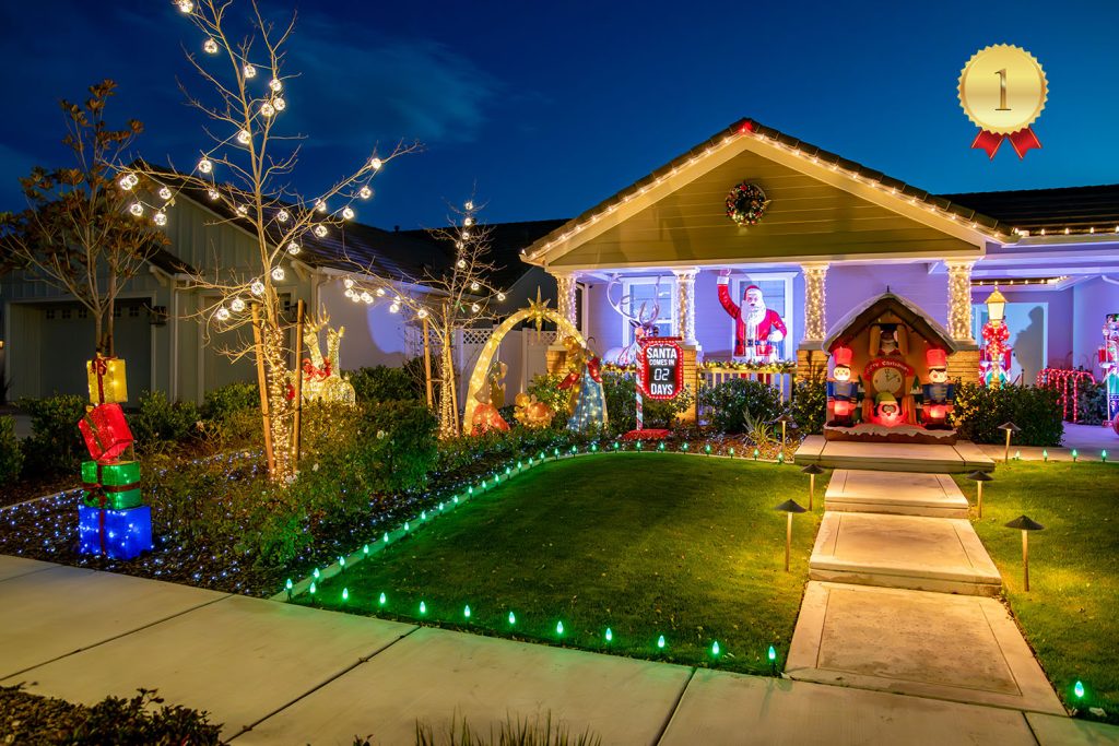 Festively decorated house at night with Christmas lights, ornaments, inflatables, and a Santa mailbox in the yard. A “1st place” badge is shown in the top right corner, indicating a holiday decorations contest winner.