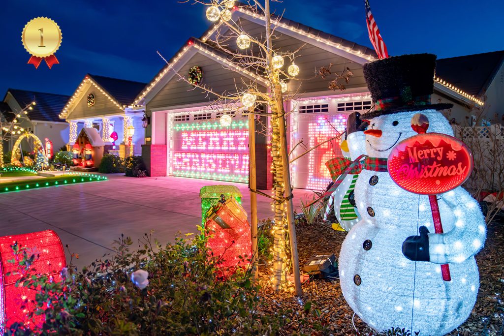 A brightly decorated house with colorful Christmas lights, a large glowing "Merry Christmas" sign, and a cheerful light-up snowman holding a "Merry Christmas" sign in the front yard at night.