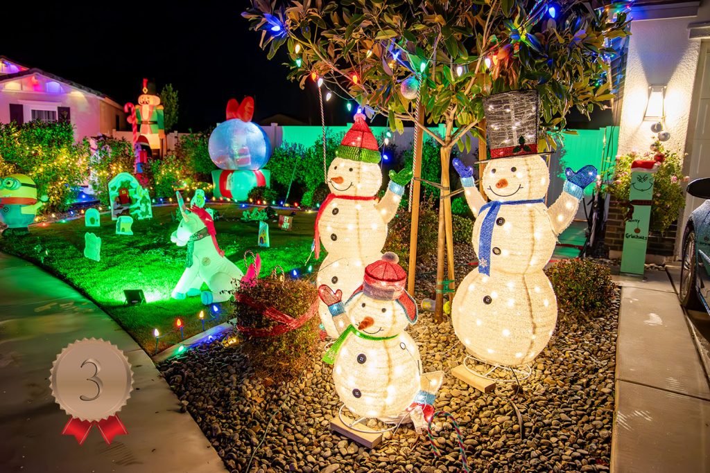 A yard decorated with glowing snowman figures, colorful holiday lights, and festive inflatables at night. Bushes and trees are lit, and a house in the background is adorned with bright seasonal decorations.