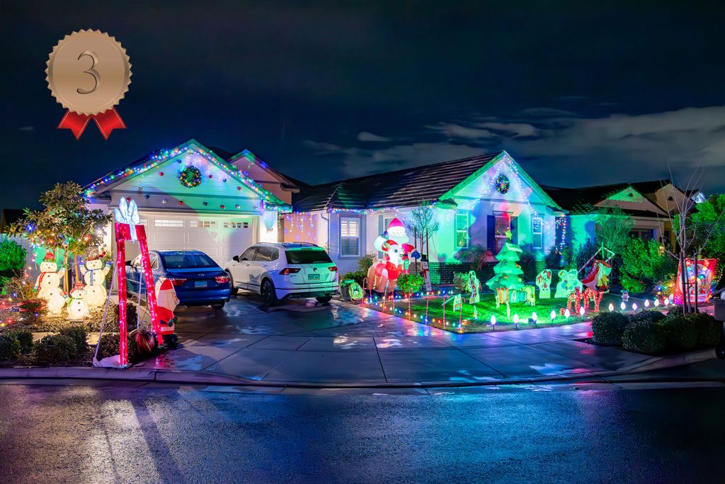 A house decorated with colorful Christmas lights, inflatable Santa and snowmen, lit reindeer, and other festive decorations. Two cars are parked in the driveway. A bronze ribbon with "3" is in the top left corner.