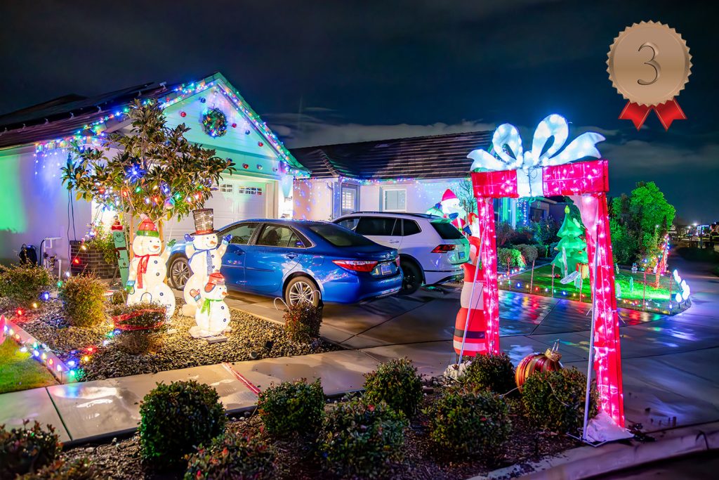 A house decorated with colorful Christmas lights, inflatable snowmen, a light-up candy cane arch, and holiday figures. Two cars are parked in the driveway. A “3rd place” ribbon is on the top right corner.