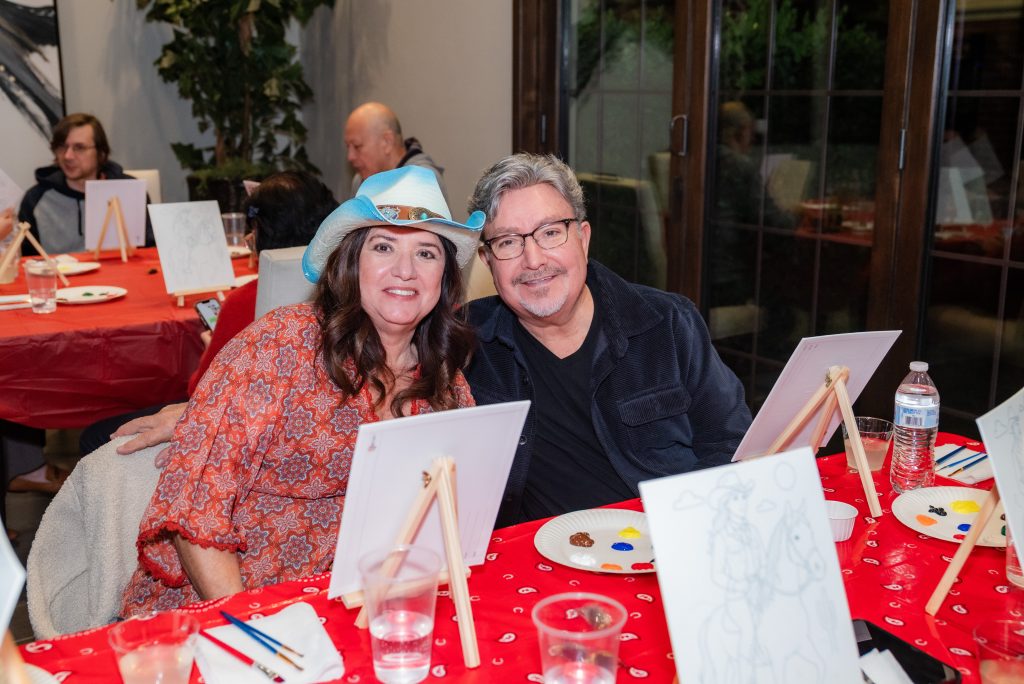 A smiling man and woman sit closely together at a table covered with a red cloth, participating in a painting activity. Blank canvases, paint, brushes, and cups of water are visible on the table. Other people paint in the background.