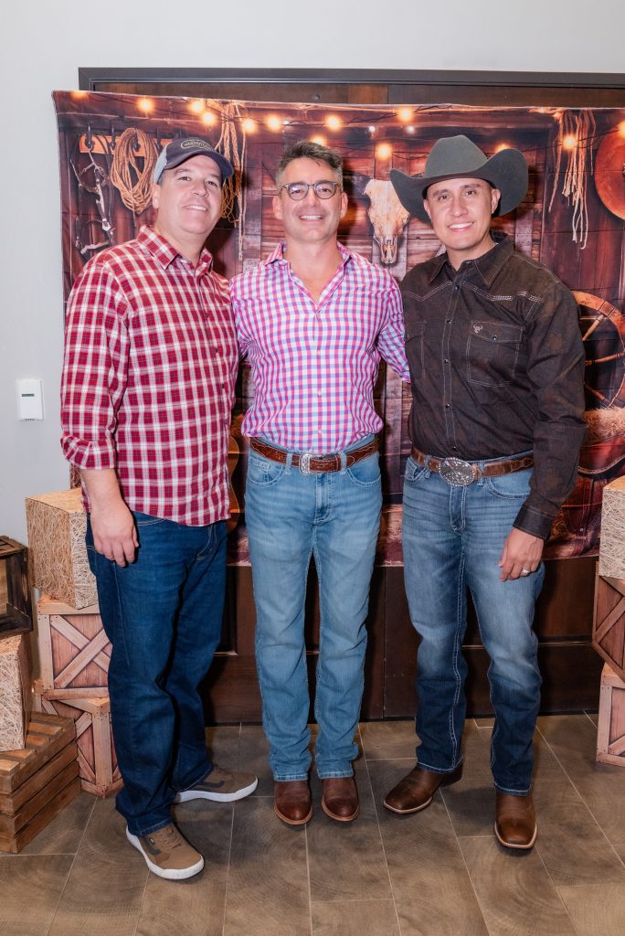 Three men stand smiling together in front of a western-themed backdrop with ropes and barrels. Two wear plaid shirts and jeans; one wears a dark shirt, jeans, and a cowboy hat. Rustic crates and wooden decor are visible.