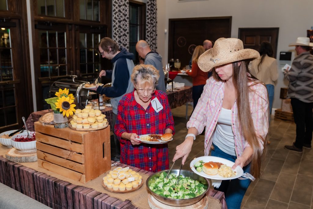 People serve themselves food from a buffet table at an indoor gathering. A woman in a cowboy hat and another in plaid are filling their plates with salad, bread, and other dishes. Cupcakes and a sunflower arrangement are on the table.