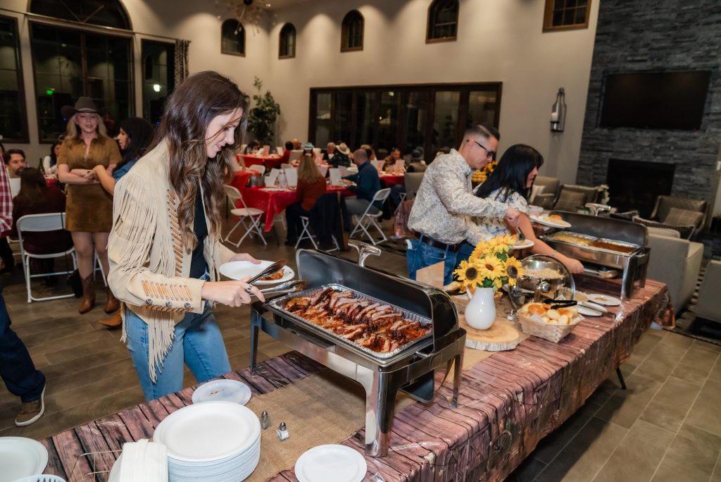 A woman serves herself barbecue from a buffet table at an indoor event, with other people in line and guests seated at round tables in the background. The buffet features chafing dishes and a centerpiece with sunflowers.