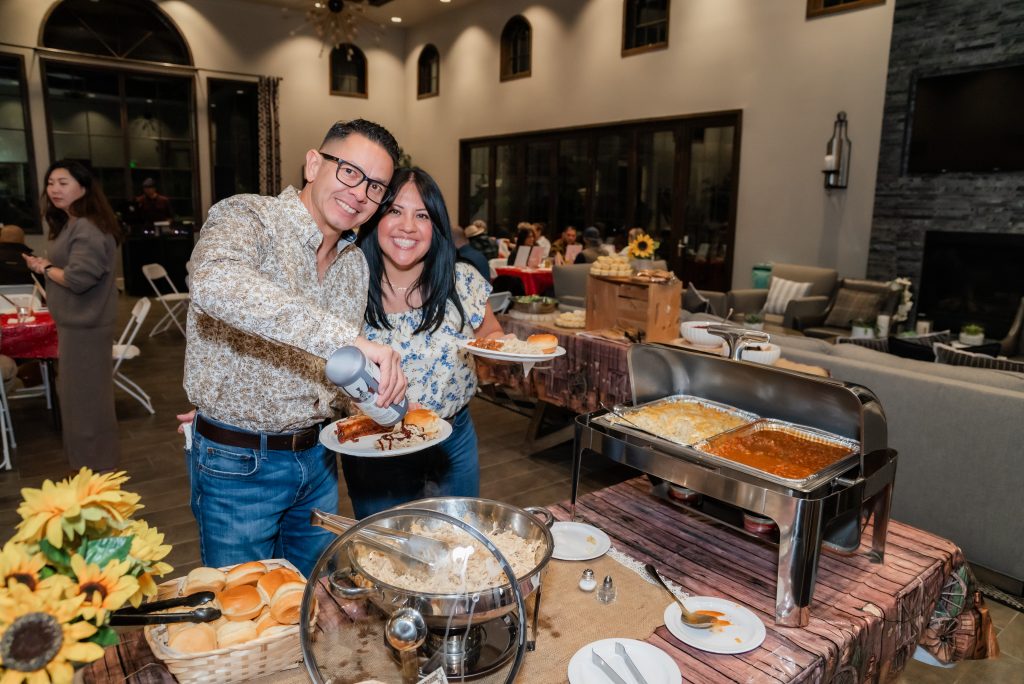 A smiling man and woman serve themselves food from a buffet at a lively indoor gathering, with chafing dishes, rolls, and flowers on the table. Guests sit and chat in the background.