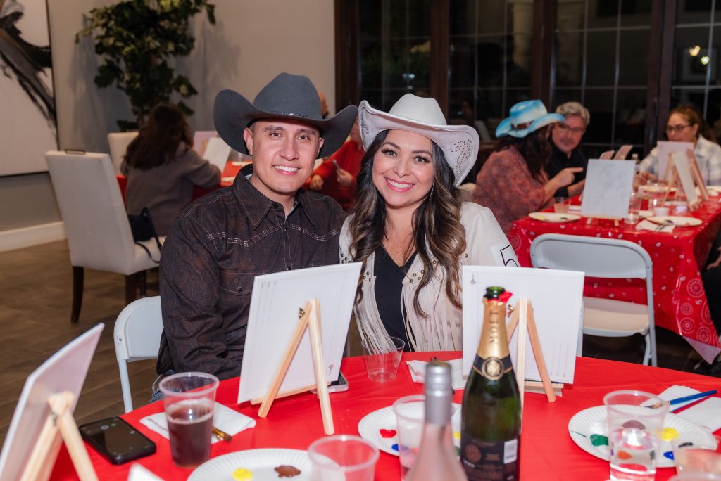 A smiling man and woman wearing cowboy hats sit at a table with painting supplies, drinks, and a champagne bottle, at a festive indoor paint party with other guests in the background.