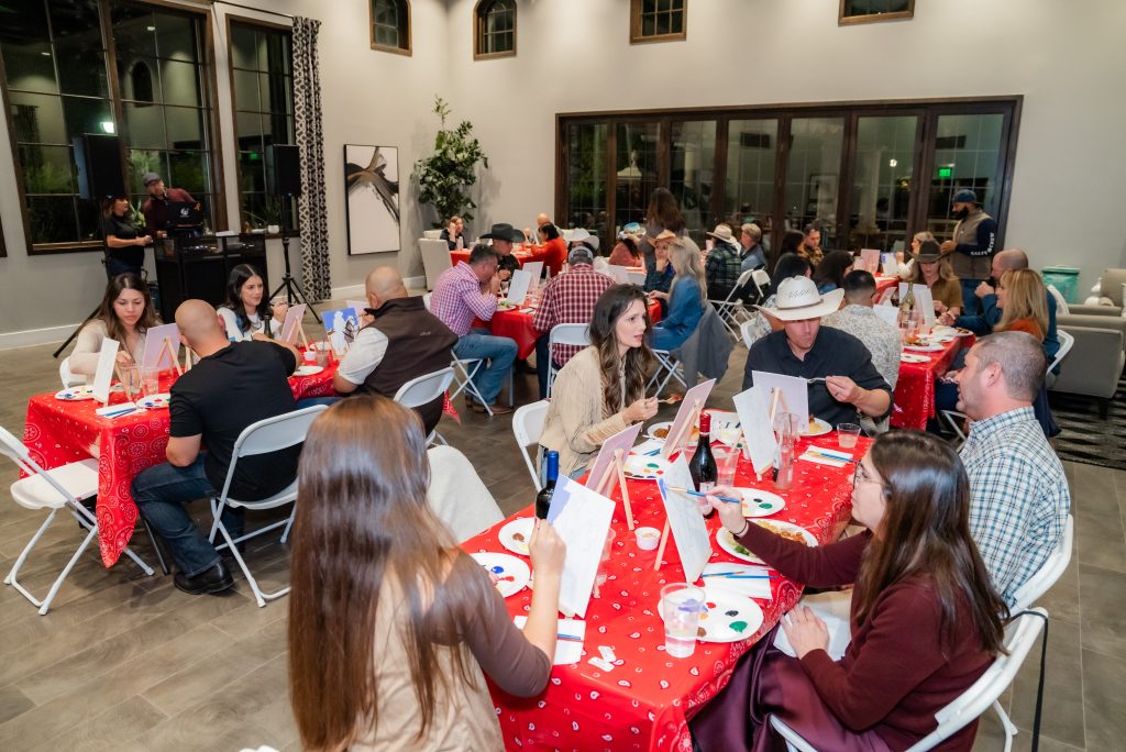 A group of people sit at tables covered with red cloths, painting canvases at a paint-and-sip event in a bright, spacious room with large windows. Art supplies and drinks are on the tables.