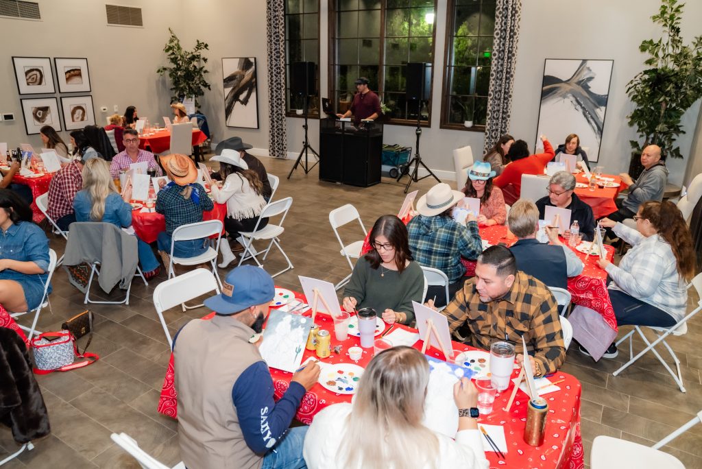 A group of people sit at round tables covered with red tablecloths, painting on canvases. Many wear cowboy hats and casual clothes. The room has framed art, large windows, and a DJ booth at the front.