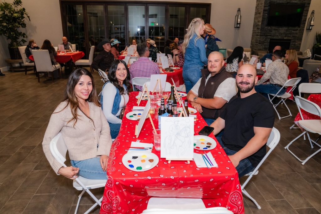 A group of four adults sit at a table covered with a red cloth, smiling at the camera during a painting event. Painting supplies and canvases are in front of them. Other groups are seen in the background.