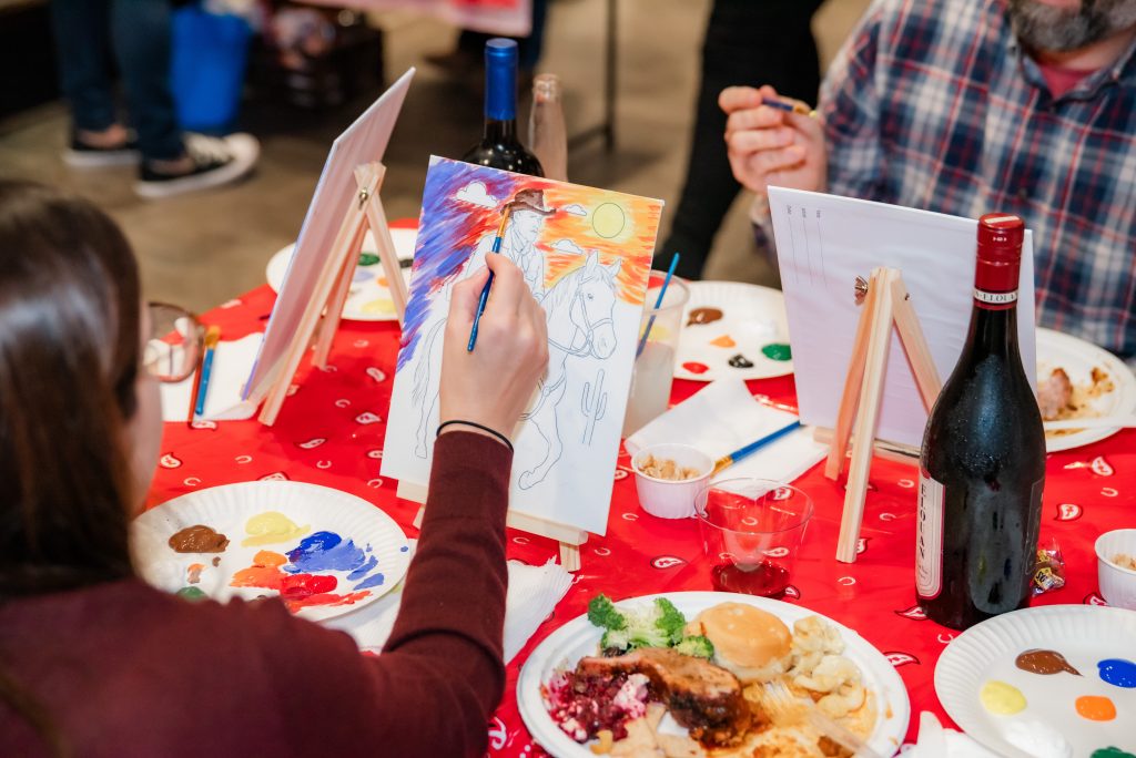 People sit around a table covered with food, drinks, and art supplies, painting at easels. One person is painting a colorful cowboy scene, and bottles of wine are visible on the table.