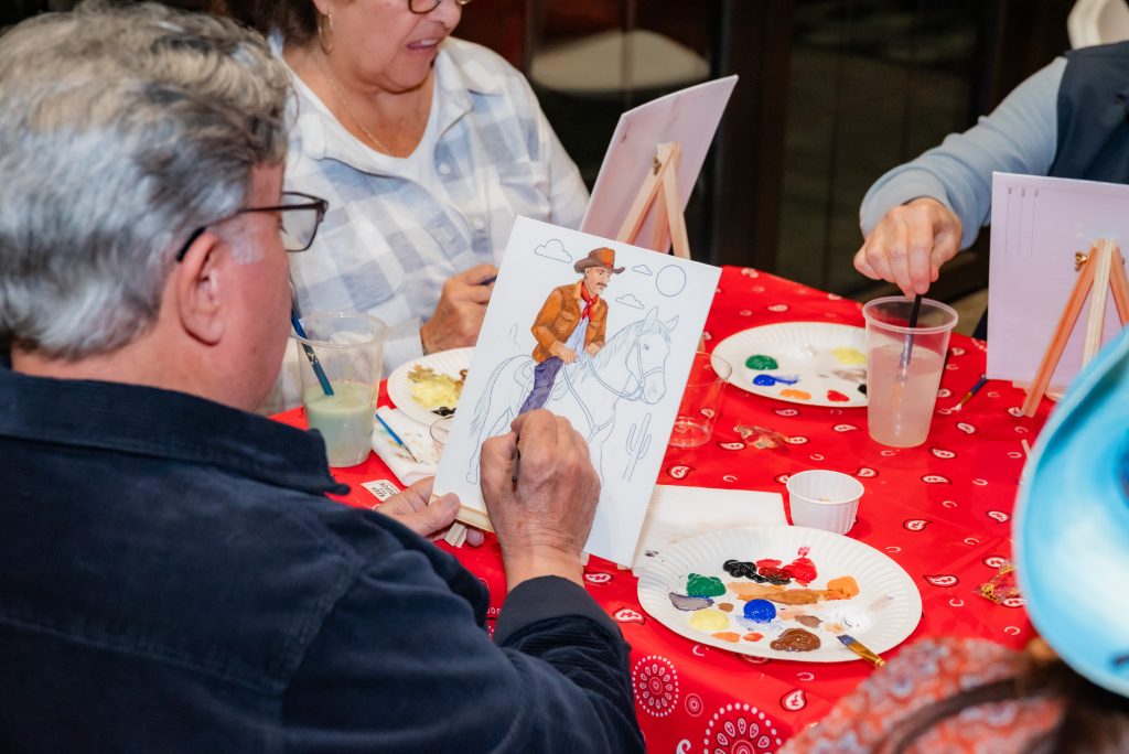 A man paints a picture of a cowboy on a horse at a table with others. Paints, brushes, and drinks are spread out on a red tablecloth with a bandana pattern.